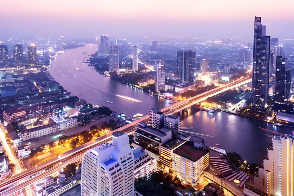 Skyline of Bangkok, Thailand at dusk, featuring the illuminated Wat Arun temple and high-rise buildings along the Chao Phraya River.